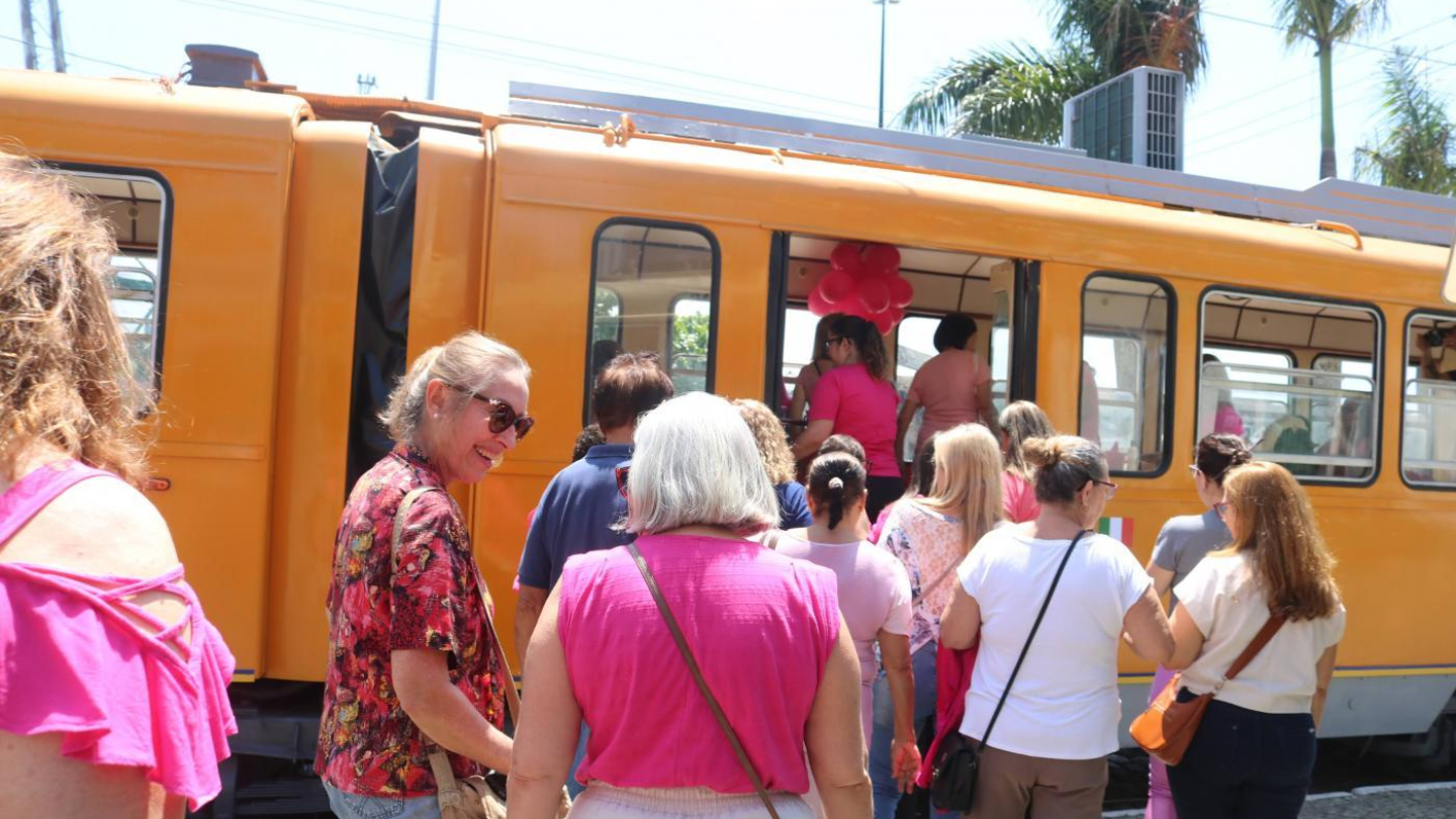 A imagem mostra um grupo de pessoas embarcando em um bonde amarelo, em um dia ensolarado. A cena transmite um clima alegre e descontraído: a maioria das pessoas veste roupas em tons de rosa, lilás e vermelho, sugerindo que participam de uma atividade temática ou comemorativa, possivelmente ligada ao Outubro Rosa, mês de conscientização sobre o câncer de mama.