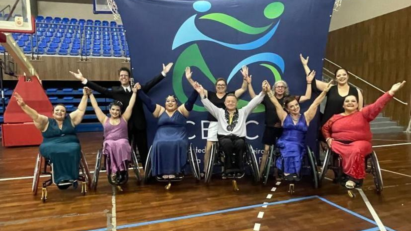 Grupo de dançarinos em cadeiras de rodas posa sorridente com os braços erguidos diante de um painel esportivo em ginásio