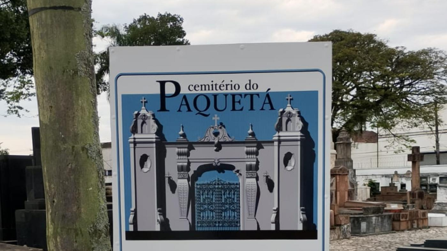 A imagem mostra uma placa informativa instalada em frente ao Cemitério do Paquetá, em Santos. A placa traz uma ilustração do portão principal do cemitério, com sua arquitetura clássica — composta por duas torres laterais, cruzes no topo e um portão ornamentado de ferro azul.  O texto na placa descreve a história do local, informando que o Cemitério do Paquetá tem uma área total de 26 mil m² e é o mais antigo da cidade, fundado em 1858. O texto menciona personalidades sepultadas ali.