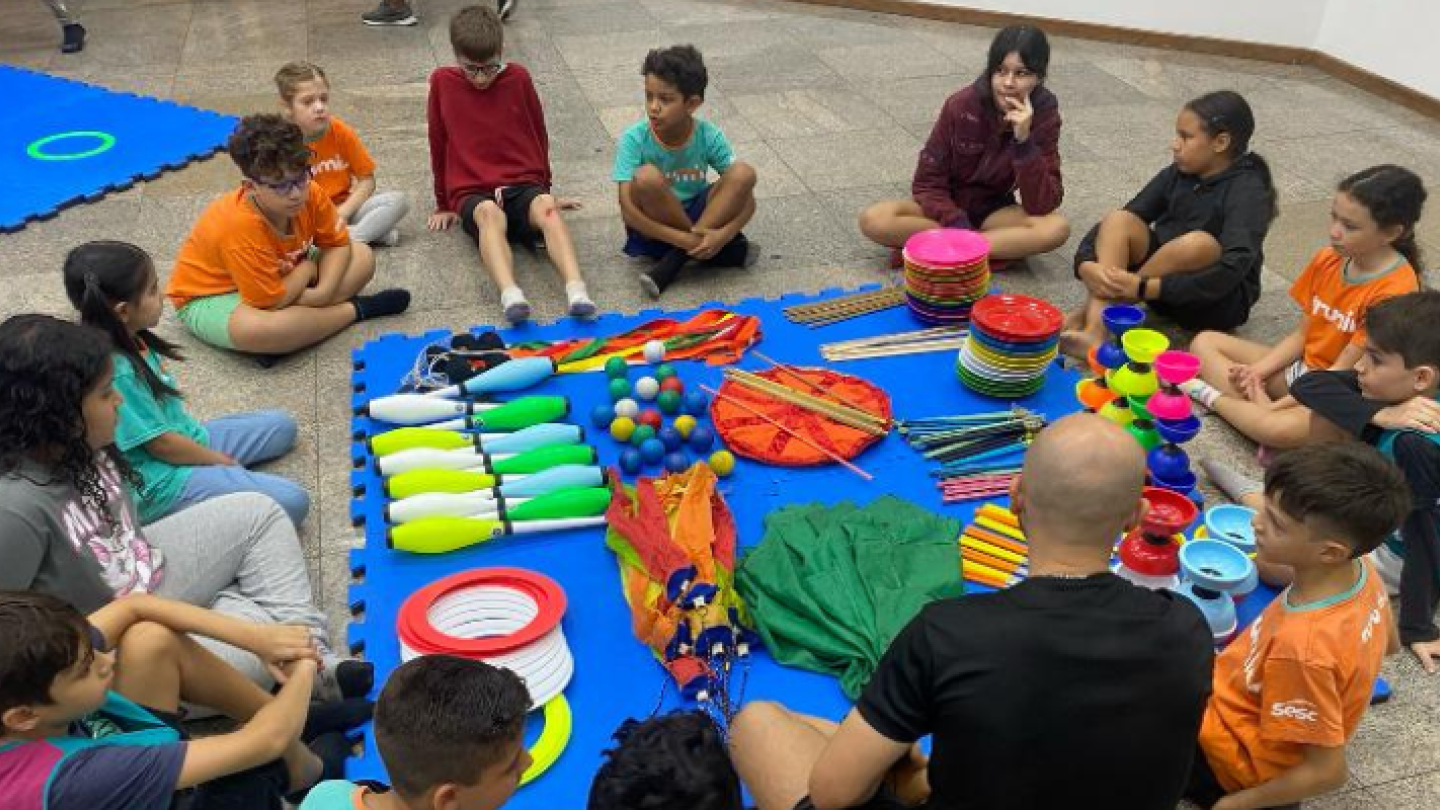 Crianças reunidas ao redor de itens coloridos de circo, como malabares, bolas, aros e tecidos