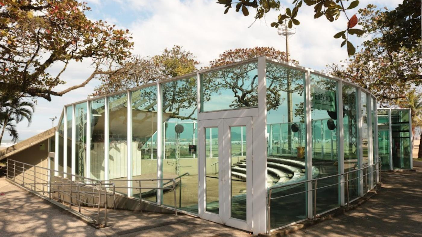 Estrutura de vidro com rampa de acesso e teto curvo, cercada por árvores, em área próxima à praia