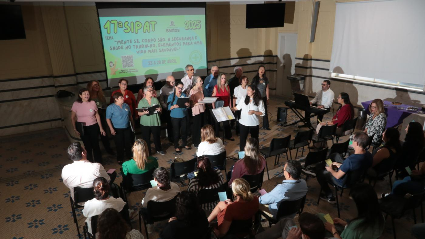 Um grupo de pessoas está em pé cantando em um coral, enquanto uma plateia sentada os observa