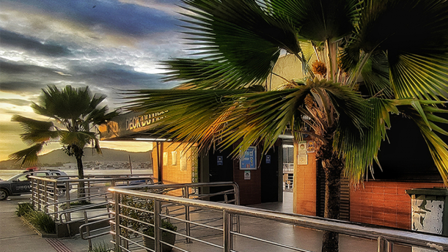 Vista do Deck do Pescador, em Santos, ao entardecer, com palmeiras, corrimãos metálicos e o mar ao fundo sob um céu parcialmente nublado