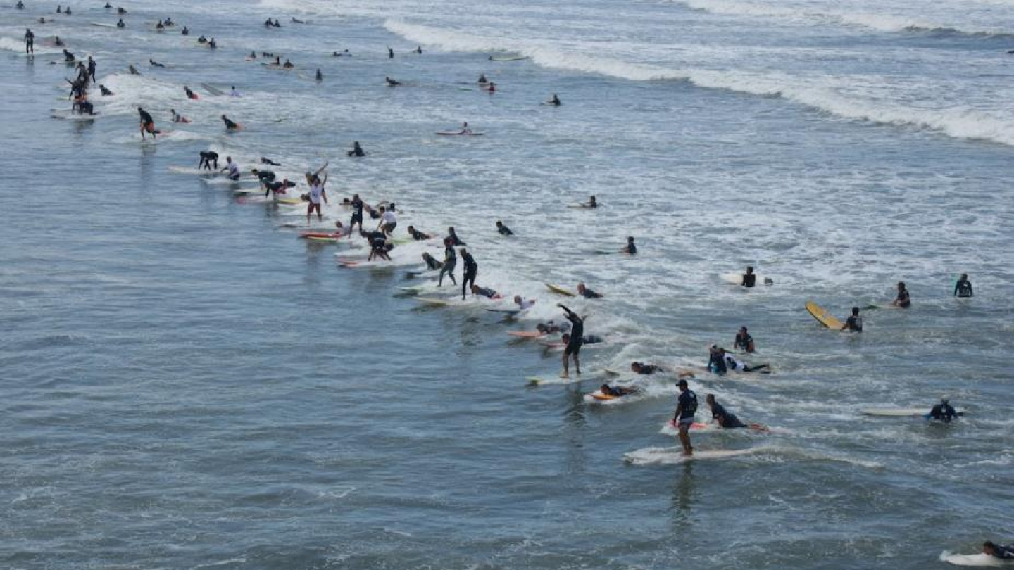 Na imagem, vemos uma cena movimentada no mar, com dezenas de surfistas pegando onda ao mesmo tempo. Muitos estão em pé nas pranchas ou se preparando para subir, e o mar está cheio de pranchas coloridas. As ondas são médias e se espalham por uma larga faixa da praia, que parece ter uma área bem ampla e rasa. O céu está claro, sugerindo um dia de sol.