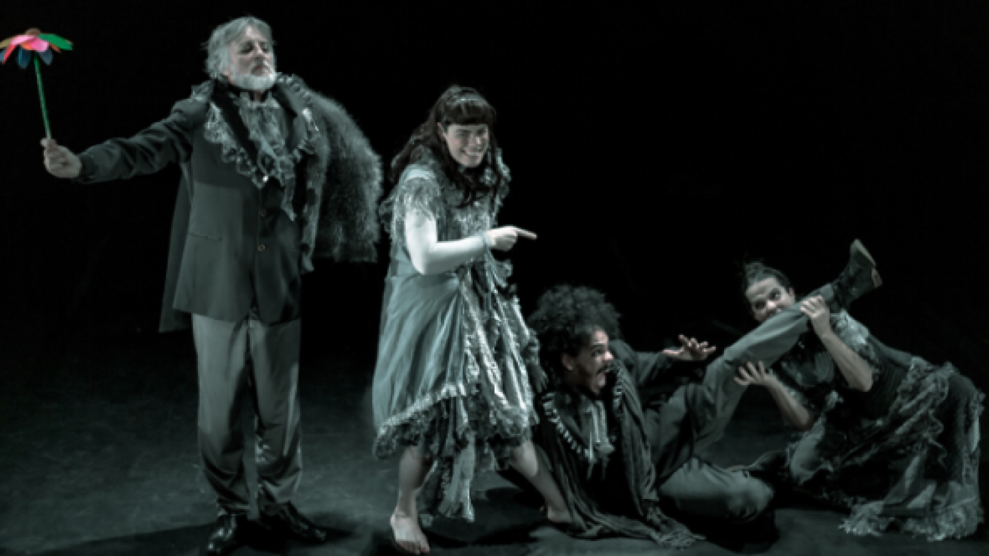 Quatro atores em cena teatral, com figurinos expressivos e gestos dramáticos, encenam momento intenso sobre palco escuro, sugerindo uma performance cômica ou simbólica