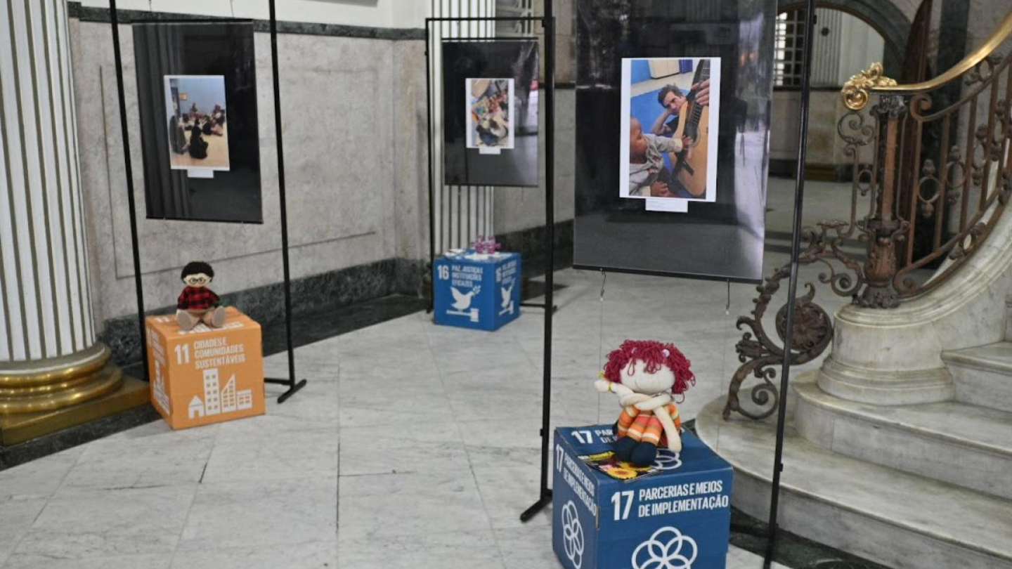 Exposição fotográfica em espaço interno com painéis pendurados e caixas coloridas dos Objetivos de Desenvolvimento Sustentável (ODS) usadas como suportes, decoradas com bonecos de pano