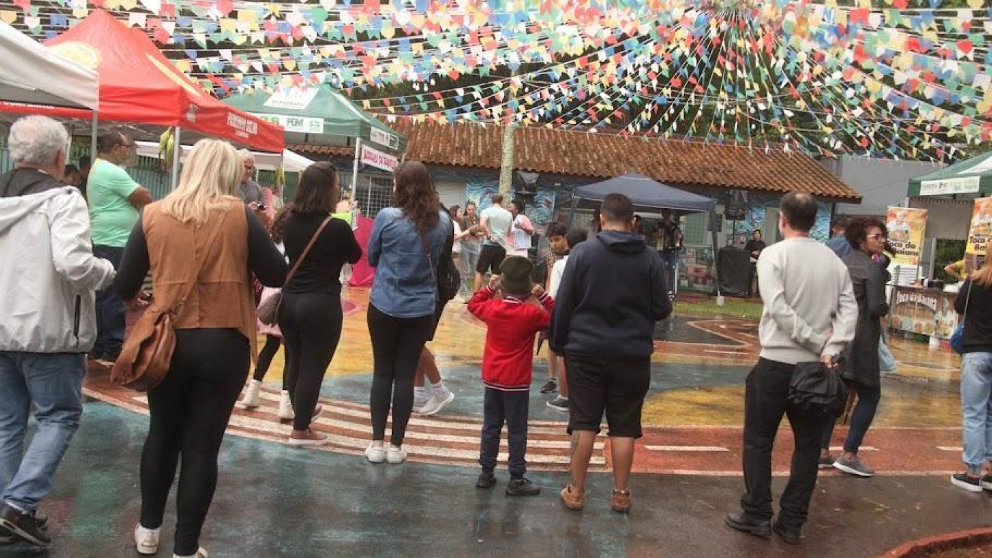 Na imagem, vemos um evento ao ar livre com clima bastante festivo. Várias pessoas estão reunidas em um espaço decorado com muitas bandeirinhas coloridas suspensas, típicas de festas populares brasileiras, especialmente festas juninas e nordestinas.  Há tendas de barracas montadas ao redor, oferecendo produtos ou comidas, com toldos em cores como vermelho, verde e branco.