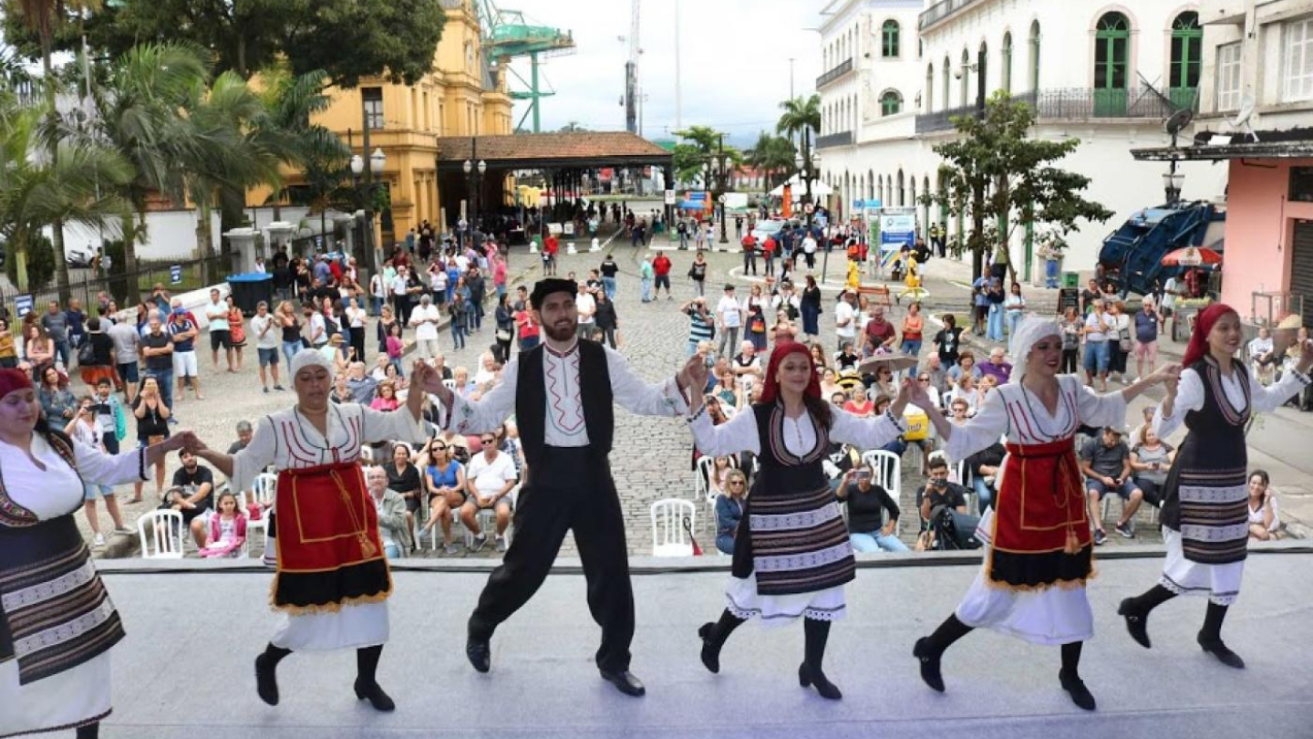 A imagem mostra uma apresentação de dança folclórica em um palco montado ao ar livre, no Largo Marquês de Monte Alegre, em frente à Estação do Valongo, em Santos. Cinco dançarinos — um homem e quatro mulheres — estão de mãos dadas, vestidos com trajes típicos gregos: as mulheres usam vestidos longos, aventais e lenços na cabeça, enquanto o homem veste calça preta, camisa branca e colete escuro. 