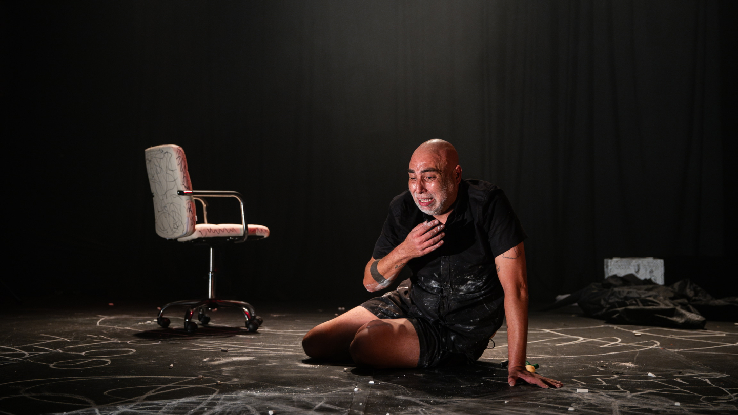 Homem se apresenta em palco escuro, ajoelhado no chão com expressão intensa, ao lado de uma cadeira