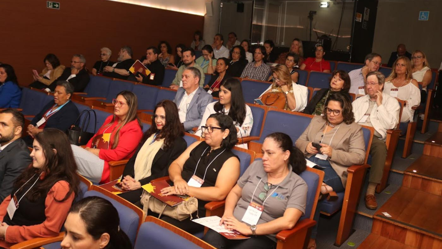 Na imagem, vê-se um auditório com diversas pessoas sentadas, acompanhando atentamente um evento. O ambiente é bem iluminado e organizado, com poltronas azuis e detalhes em madeira. A plateia é composta por homens e mulheres de diferentes idades, muitos usando crachás de identificação, o que indica tratar-se de um encontro formal, como um fórum, congresso ou seminário.