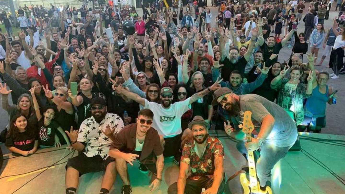 A imagem registra um grande grupo de pessoas reunidas em um evento ao ar livre, com clima festivo e descontraído. Em primeiro plano, sobre o palco, membros de uma banda posam sorrindo para a foto. Um dos músicos segura um contrabaixo, enquanto os demais fazem gestos de animação e cumprem a função de animadores do público. Todos parecem satisfeitos com a apresentação.