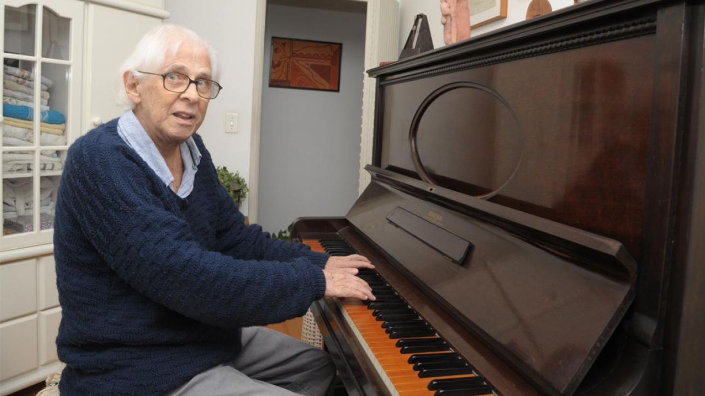 Na imagem, vemos um homem idoso sentado ao piano, em um ambiente interno que parece ser uma sala ou escritório. Ele tem cabelos brancos, usa óculos, um suéter azul e calças claras. Suas mãos estão posicionadas sobre as teclas do piano, como se estivesse prestes a tocar ou já tocando. Ao fundo, há móveis claros com toalhas dobradas e um quadro decorativo na parede.