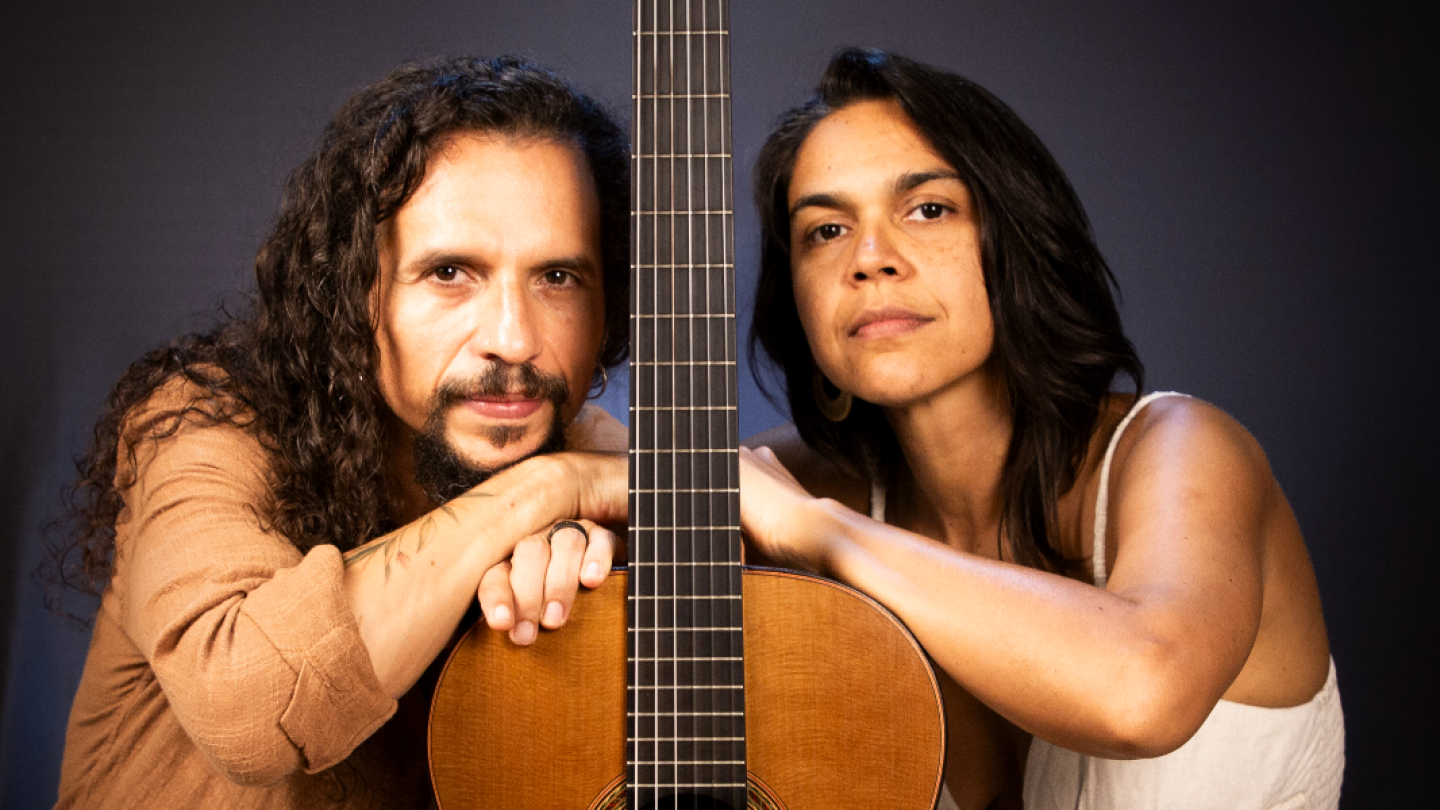 Homem e mulher apoiados num violão, com fundo escuro