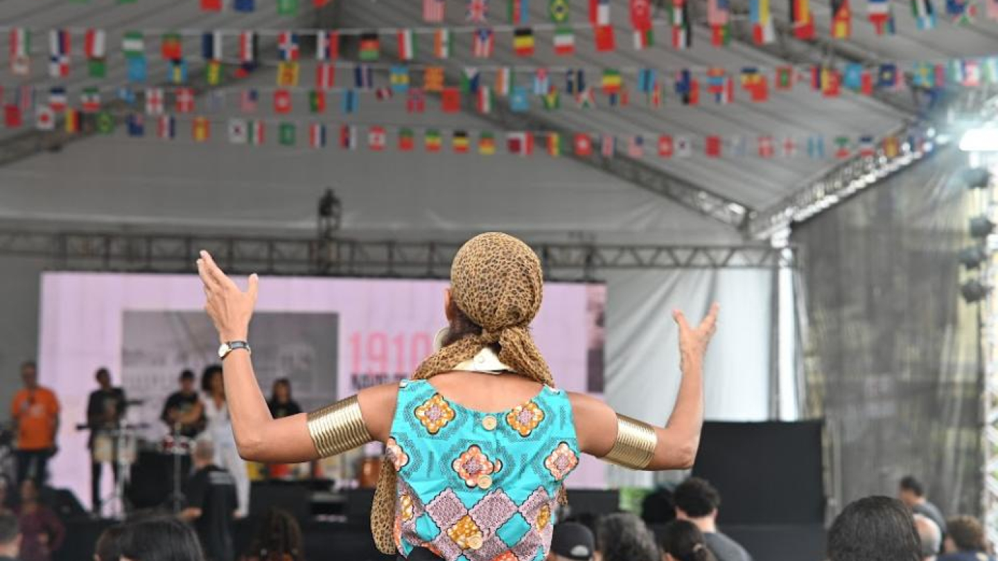 Ela mostra um momento vibrante do Festival do Imigrante, realizado sob uma grande tenda decorada com bandeiras de diversos países penduradas no teto, simbolizando a diversidade cultural presente no evento. Em primeiro plano, uma pessoa aparece de costas, com os braços erguidos em gesto expressivo, vestindo roupas coloridas com estampas geométricas e acessórios dourados nos braços e na cabeça. À frente, o palco abriga músicos e técnicos, enquanto o público, sentado, acompanha atentamente a apresentação.
