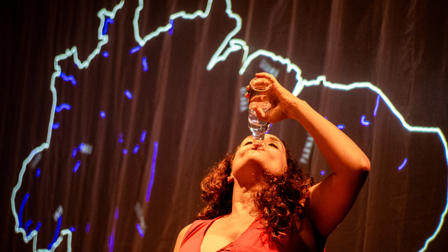 Mulher de roupa vermelha bebe de um copo transparente diante de um fundo iluminado com o contorno brilhante do mapa do Brasil, criando uma cena artística e teatral