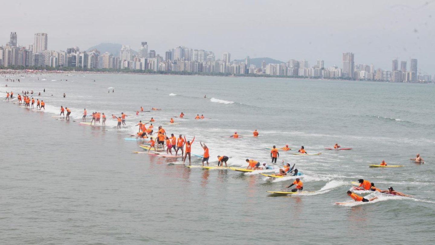 A imagem mostra um grande grupo de surfistas no mar, participando de um evento coletivo em uma praia urbana. Todos vestem camisetas laranja, o que dá um visual vibrante e uniforme à cena. Muitos estão de pé nas pranchas, outros remam ou se equilibram nas ondas pequenas e rasas.