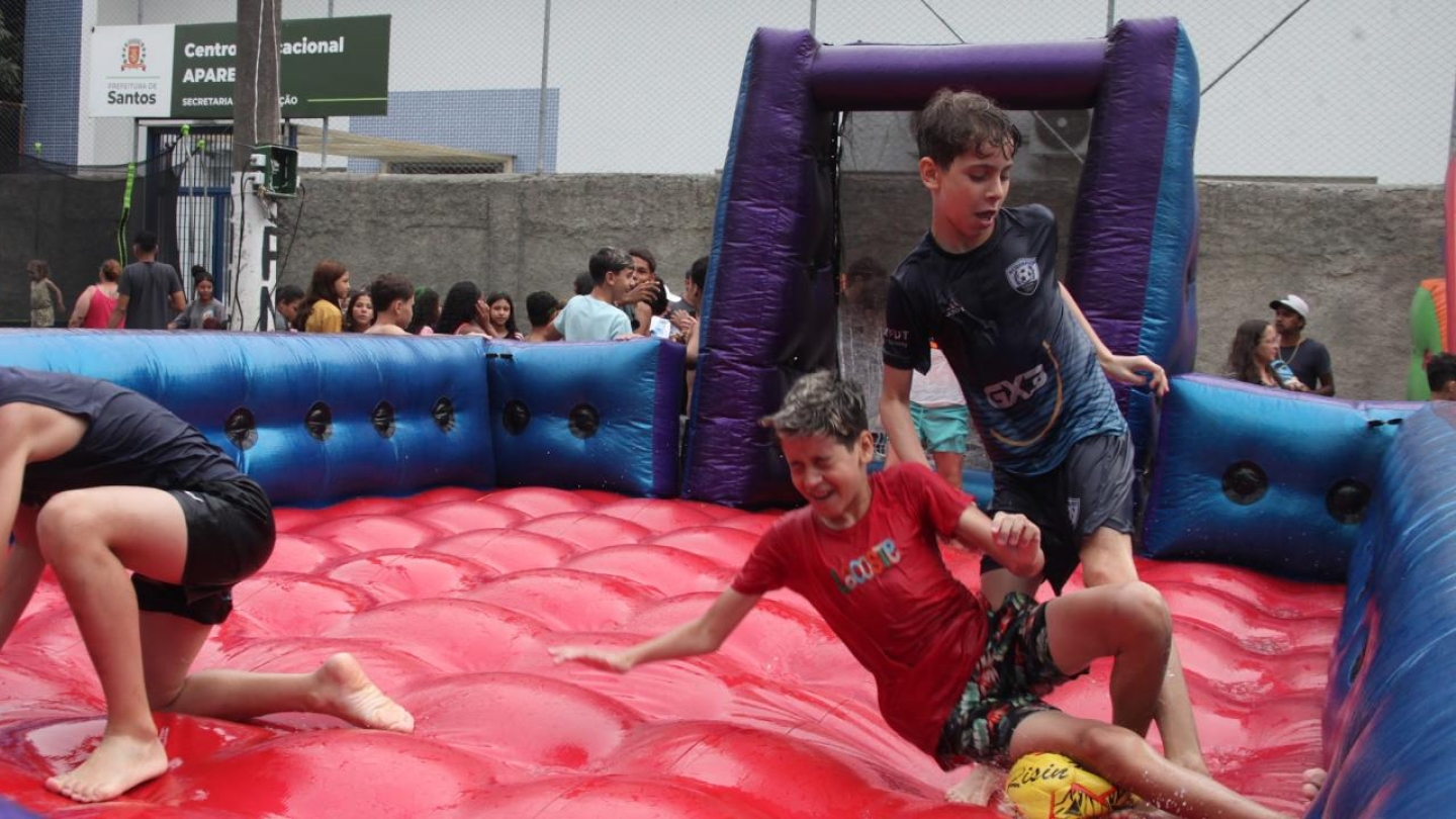 A imagem mostra três meninos brincando em um brinquedo inflável molhado, com chão vermelho e laterais azuis, em uma atividade ao ar livre. Eles parecem se divertir bastante, escorregando e rindo enquanto tentam manter o equilíbrio. Ao fundo, há outras crianças e adultos observando, e uma placa indica que o local é o Centro Esportivo e Recreativo do Aparecida, da Prefeitura de Santos.