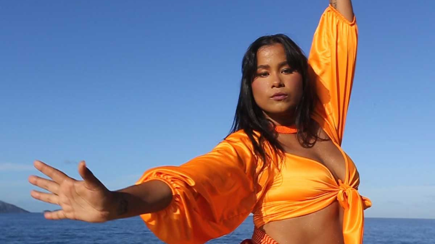 Mulher, de traços indígenas, com braços abertos e traje amarelo