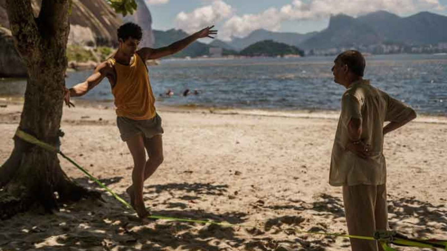 Homem pratica slackline na praia enquanto outro observa, com o mar e montanhas ao fundo em um dia ensolarado