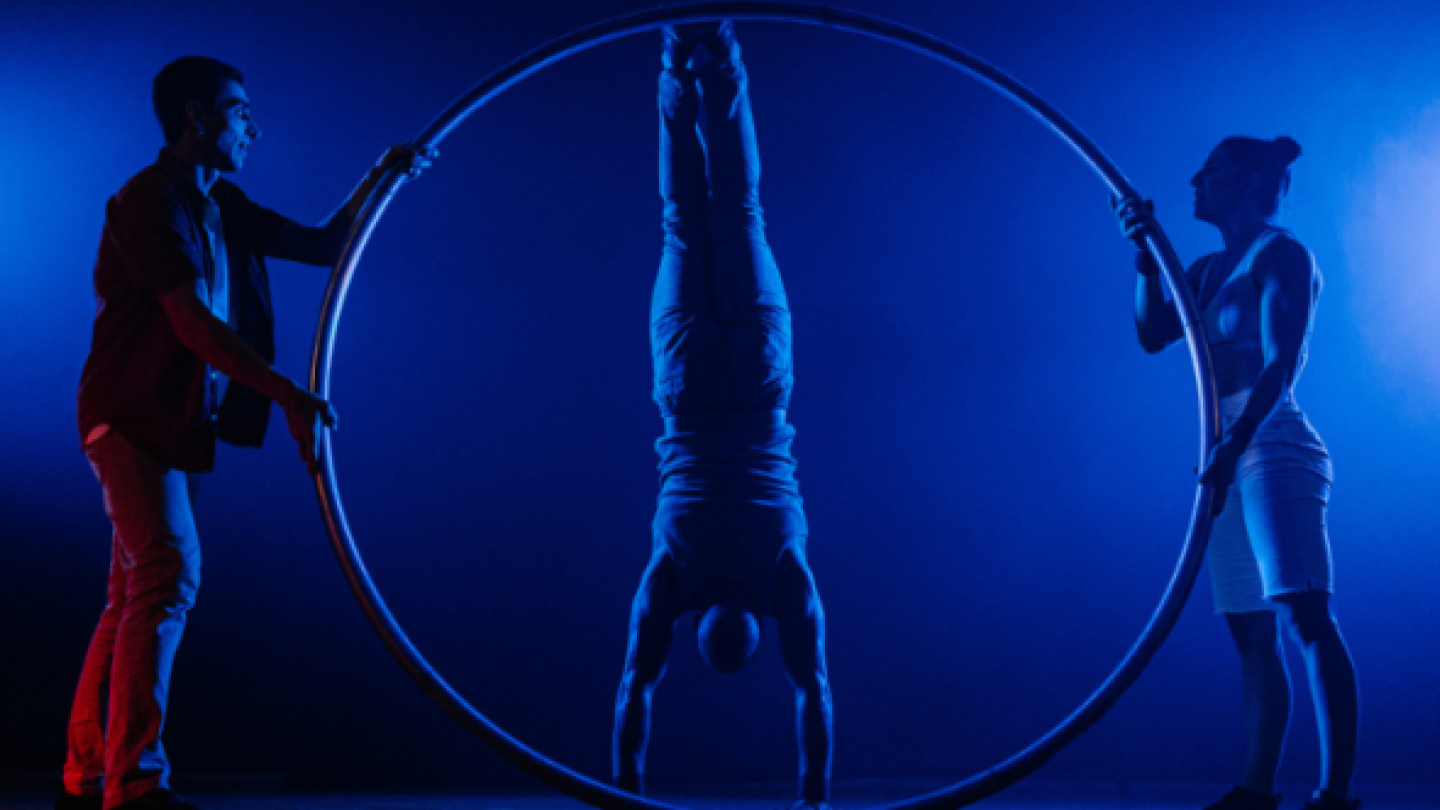 Três artistas circenses se apresentam sob luz azul; um realiza um equilíbrio de ponta-cabeça dentro de um aro metálico, enquanto os outros dois o sustentam