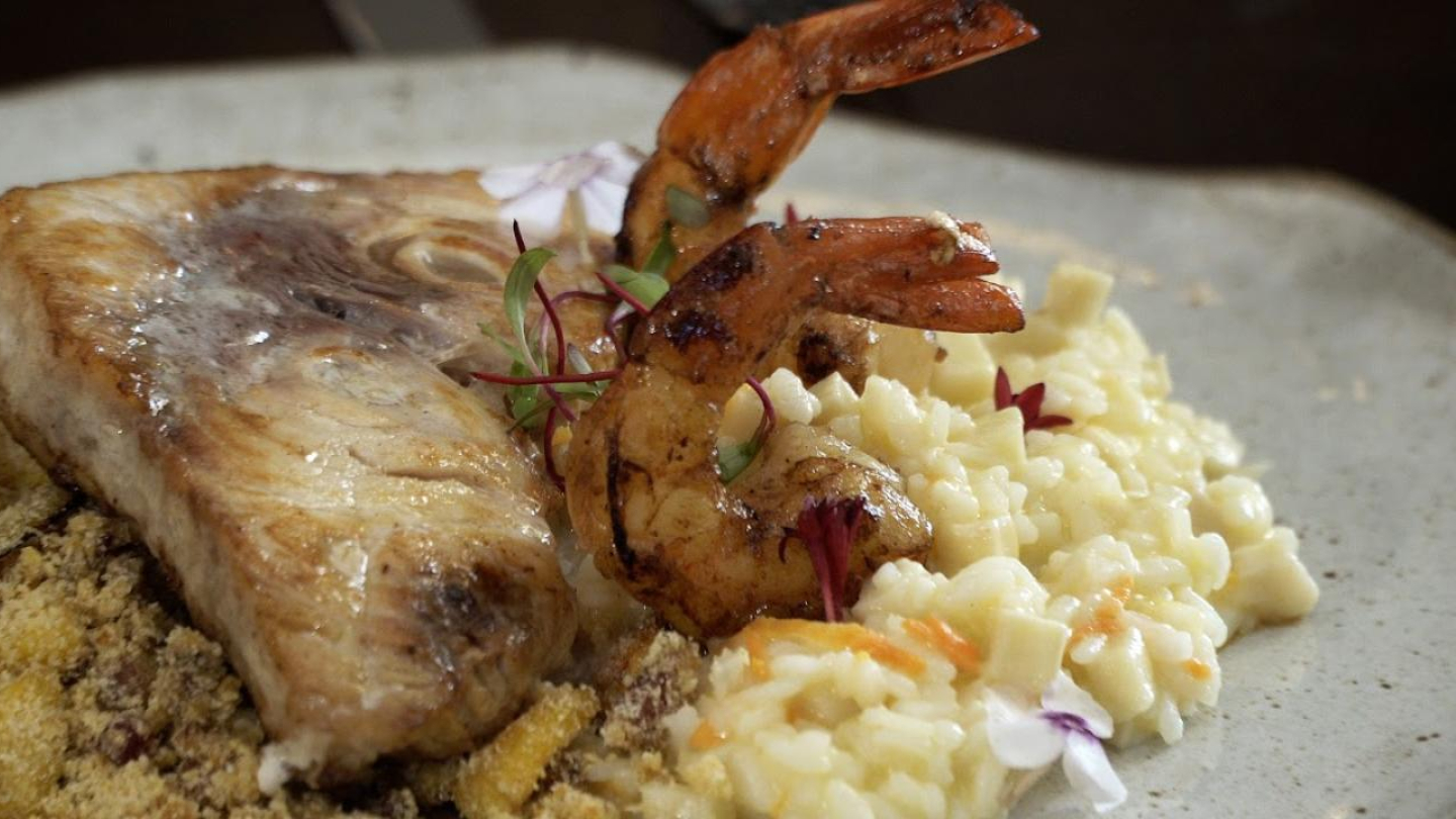 Na imagem, vemos um prato sofisticado de frutos do mar. No centro, há uma posta de peixe grelhado, de aparência suculenta e dourada, servida sobre uma farofa crocante. Ao lado, há dois camarões grelhados com leve brilho, indicando que foram preparados na manteiga ou azeite. O acompanhamento parece ser um risoto cremoso, possivelmente de pupunha ou legumes, decorado com pequenas flores comestíveis e brotos, que dão um toque delicado e colorido ao prato. A montagem é elegante e apetitosa.