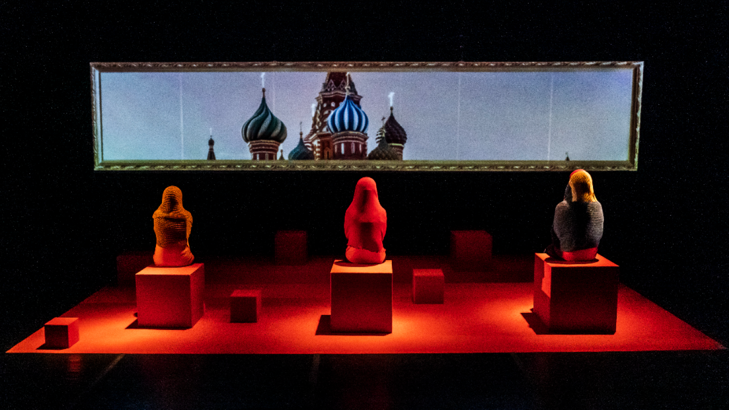 Uma cena teatral iluminada por luz vermelha no chão, com três figuras sentadas de costas em pedestais cúbicos vermelhos e, ao fundo, uma tela com imagem da Catedral de São Basílio em Moscou