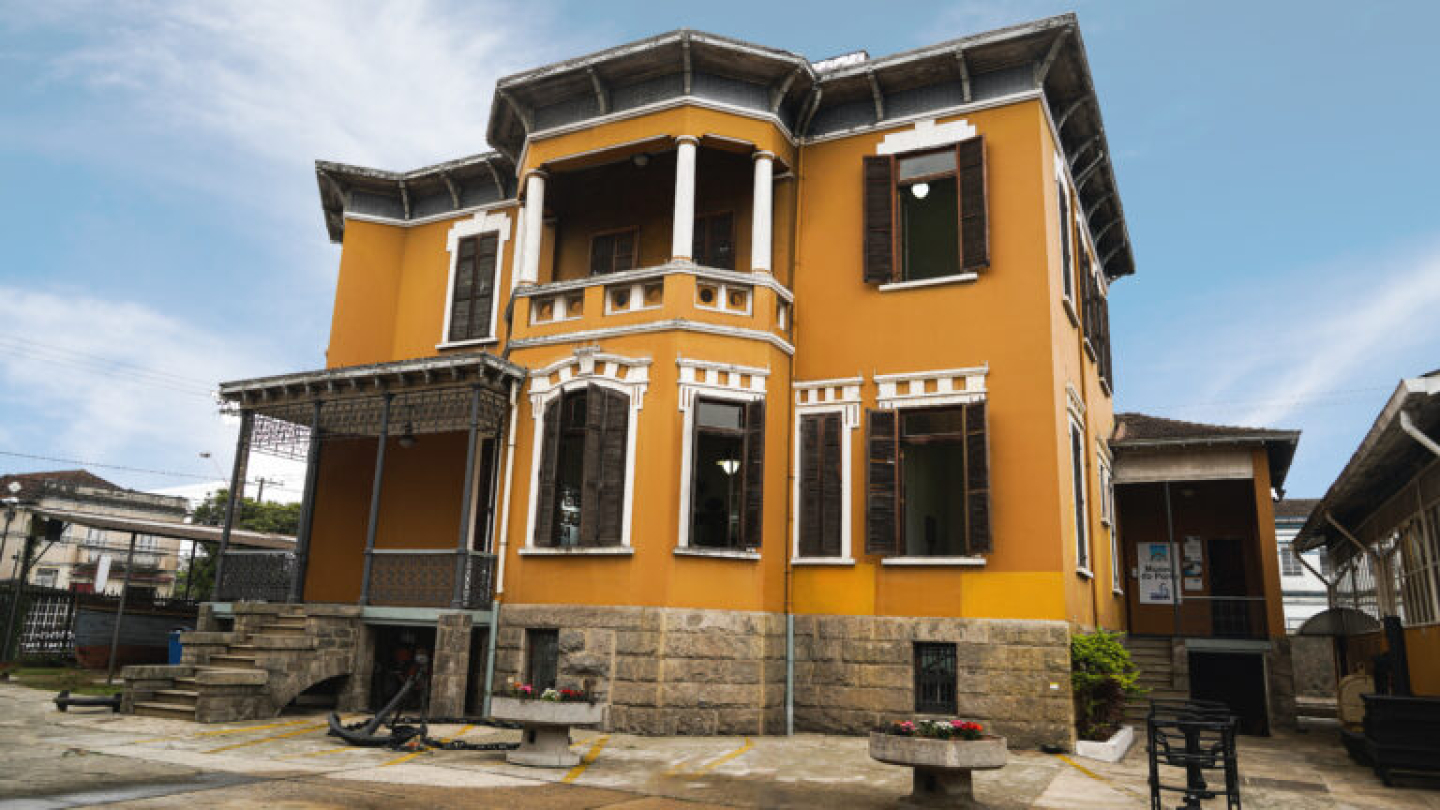 Prédio histórico de dois andares com fachada em tom ocre e detalhes brancos, janelas com venezianas escuras e varanda com colunas brancas. A base da construção é de pedra, e há um pátio com bancos e canteiros de flores ao redor