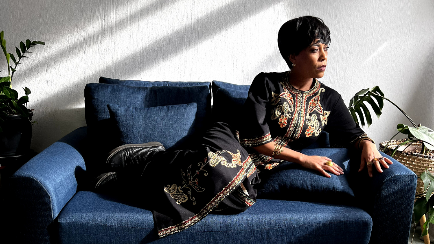 Mulher sentada em um sofá azul, vestindo roupa preta com bordados dourados e coloridos, olha pensativa para o lado sob a luz natural