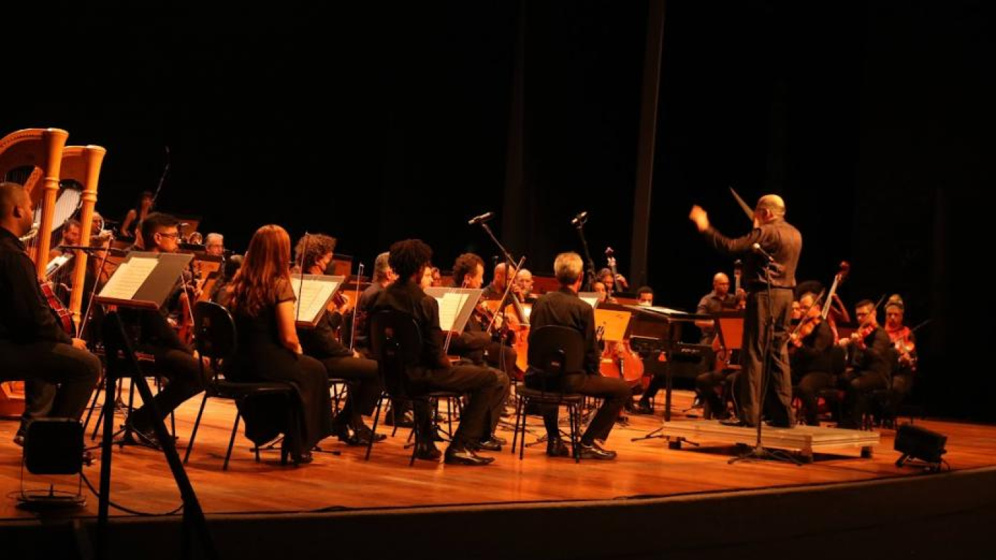 Apresentação de Orquestra Sinfônica, com todos de preto e maestro liderando à frente
