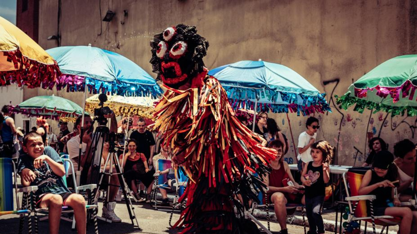 Pessoa com fantasia colorida e máscara grande se apresenta em rua decorada com guarda-sóis, diante de um público animado