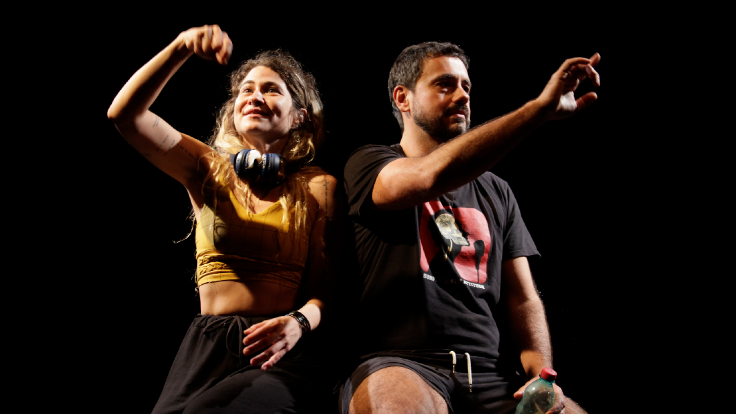 Uma mulher e um homem, vestindo roupas casuais, estão sentados em um palco escuro, ambos com os braços levantados e apontando ou gesticulando com as mãos em uma performance teatral intensa