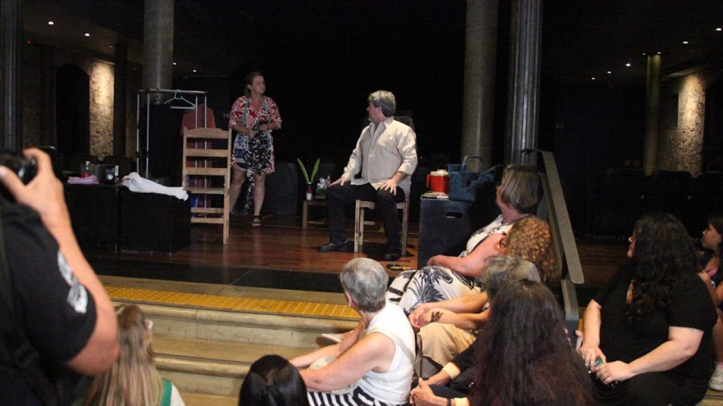 A imagem mostra uma cena teatral acontecendo em um palco. Há dois atores em destaque: uma mulher em pé, vestindo um vestido estampado, e um homem sentado em uma cadeira, usando camisa clara e calça escura. Eles parecem estar encenando um diálogo.  Na frente do palco, há um pequeno público sentado nos degraus, assistindo atentamente à apresentação — composto por mulheres e algumas crianças. À esquerda, uma pessoa com camiseta preta e mochila fotografa a cena.
