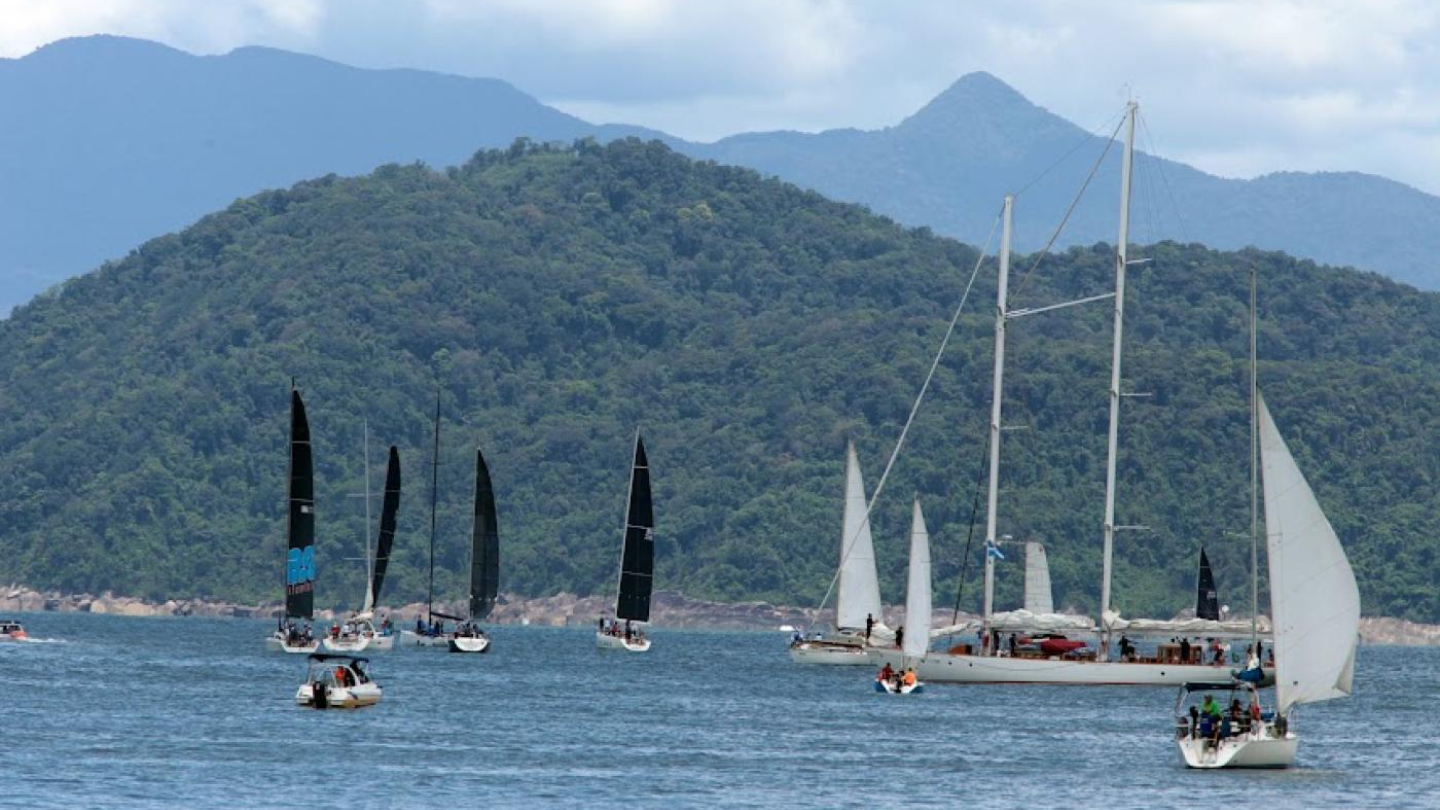 Uma foto de uma regata, mostrando diversos veleiros com velas brancas e pretas navegando no mar, com montanhas ao fundo