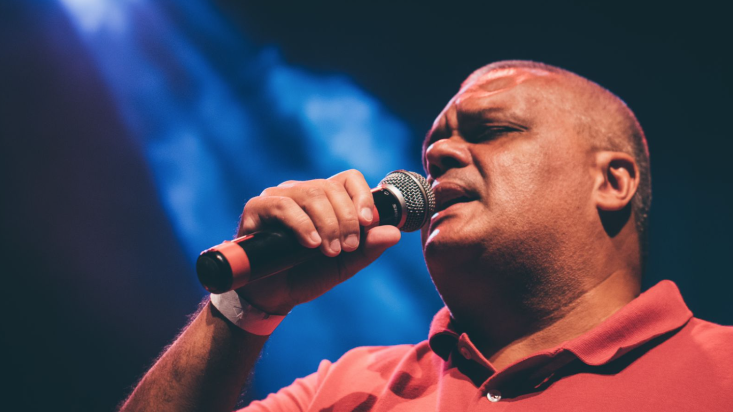 Homem trajando uma camisa polo vermelha, segura microfone, com expressão concentrada, sob uma luz azul
