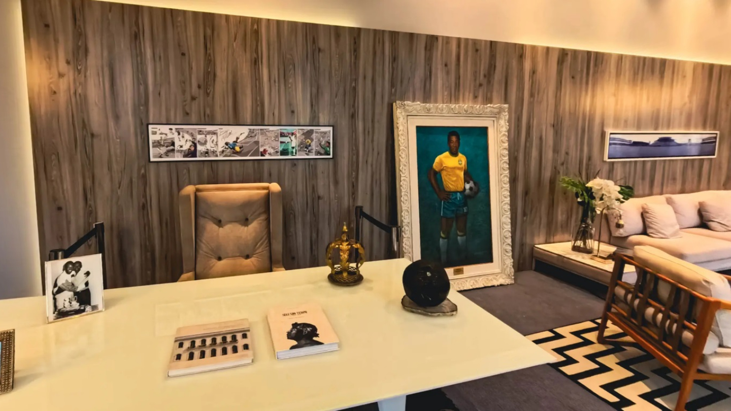 A imagem mostra um ambiente elegante e bem iluminado, que parece ser um escritório ou sala de homenagens. O destaque é um grande retrato emoldurado de um jogador de futebol vestindo o uniforme da Seleção Brasileira, com uma bola apoiada ao lado — a pintura está disposta no chão, encostada em uma parede de madeira clara.