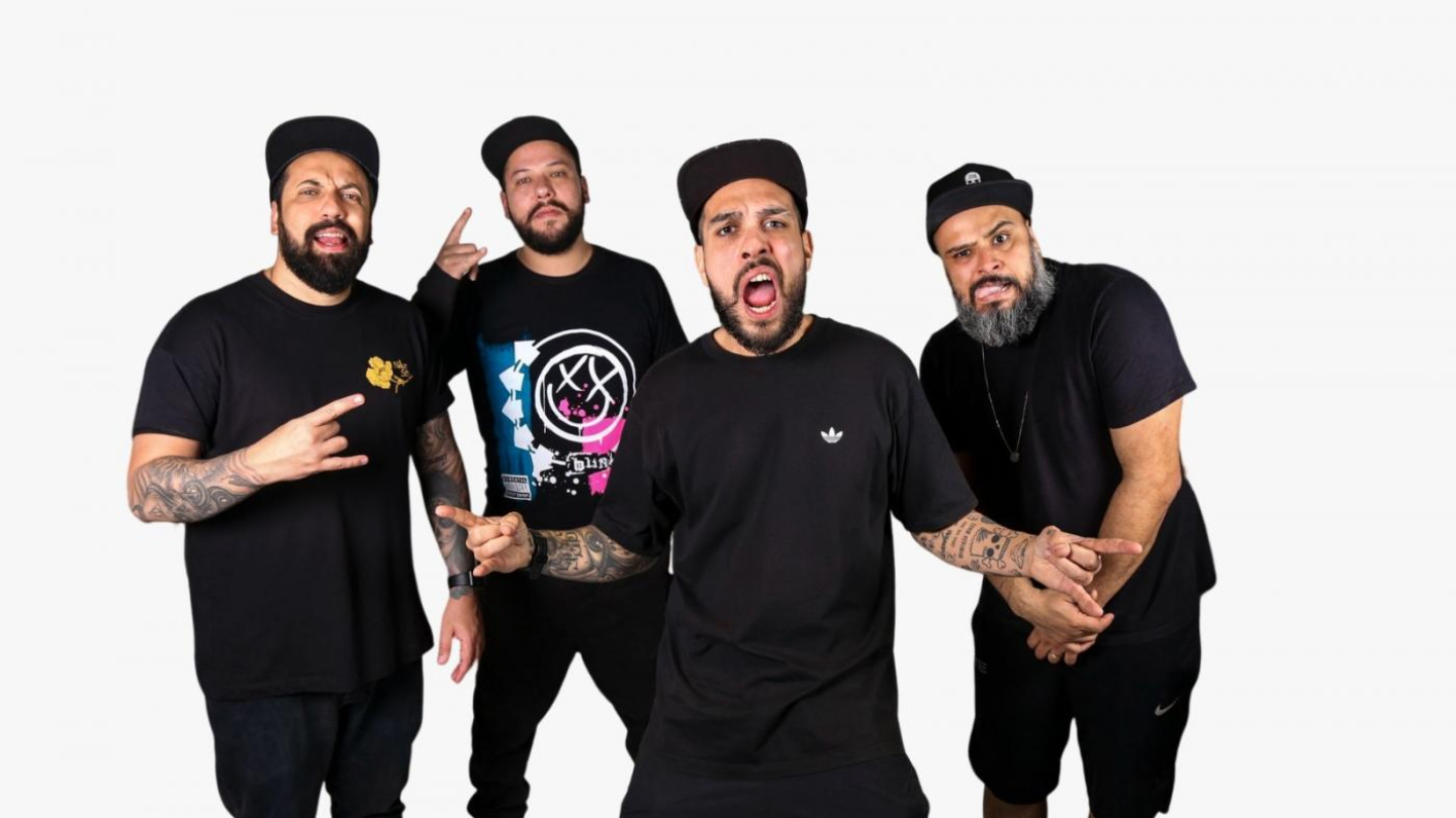 A imagem mostra quatro homens posando de forma descontraída diante de um fundo branco. Todos usam roupas escuras — majoritariamente pretas — e bonés. Eles exibem expressões animadas e gestos típicos de quem está se divertindo, como mãos fazendo sinais de “rock” ou apontando para a câmera.