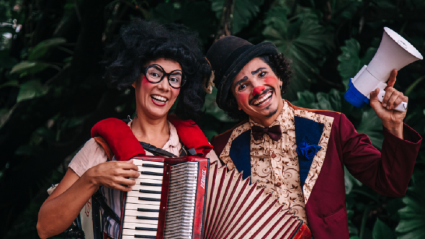 Dois artistas caracterizados como palhaços sorriem; um toca sanfona e o outro segura um megafone, em cenário com vegetação ao fundo