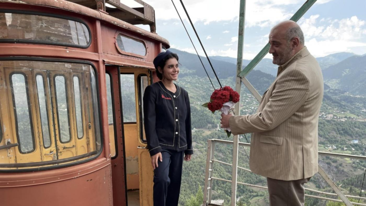 A imagem mostra uma cena capturada em um ponto de teleférico, em uma região montanhosa. À esquerda, aparece uma cabine antiga de teleférico, em tons de vermelho e amarelo, com janelas arredondadas e sinais de desgaste pelo tempo.  Diante da cabine, está uma mulher vestida com uniforme azul-escuro, calçando botas pretas e usando um boné. Ela sorri levemente, olhando para um homem à sua frente. 