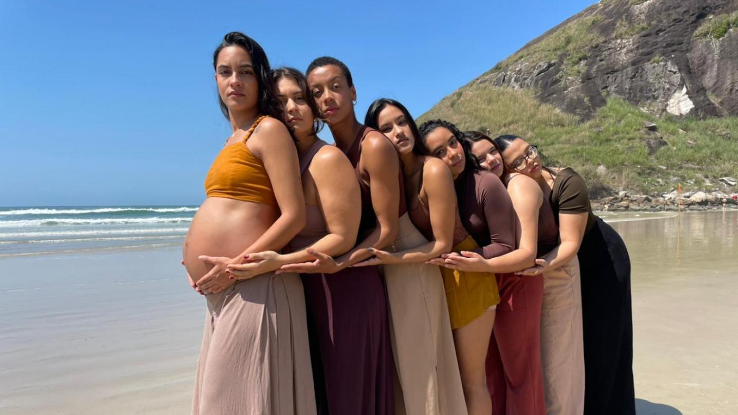 Na imagem, há sete mulheres alinhadas em uma praia, formando uma composição harmoniosa e simbólica. Todas estão descalças sobre a areia molhada, e o mar, com pequenas ondas, aparece ao fundo sob um céu azul. A mulher à frente está grávida e segura a barriga com as mãos, enquanto as demais a abraçam em sequência, apoiando as mãos sobre a barriga dela — um gesto de união, acolhimento e conexão feminina.  Elas vestem saias e tops ou blusas em tons terrosos e neutros (como marrom, bege, vinho e mostarda).