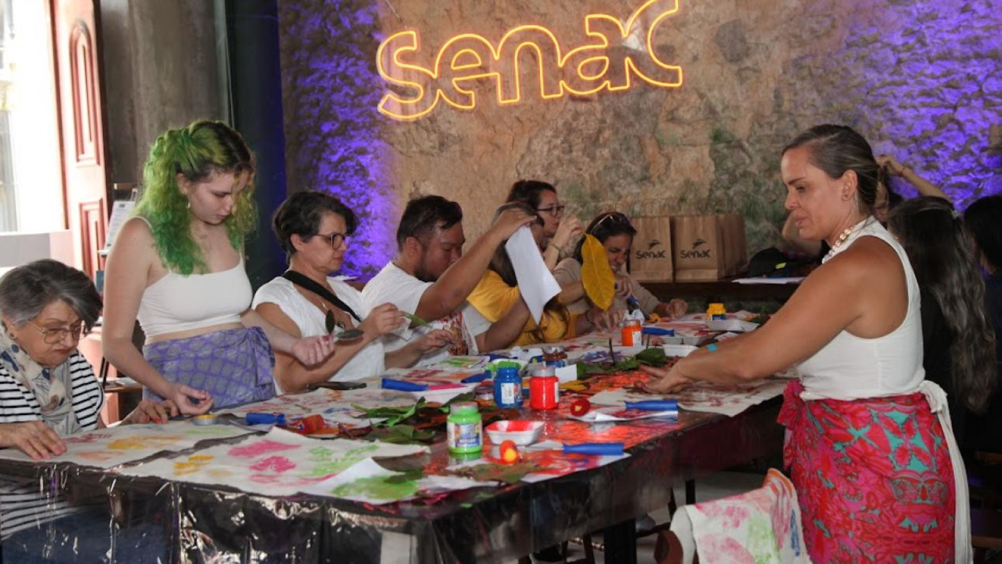 Na imagem, vemos um grupo de pessoas participando de uma atividade artística em uma mesa comprida, coberta com plástico protetor e materiais de arte como tintas coloridas, pincéis, tesouras, folhas e papéis. A ação acontece em um ambiente interno com paredes de pedra aparentes e iluminação decorativa em tons roxos.  Ao fundo, há uma placa iluminada com o nome “Senac”, indicando que a atividade está sendo realizada em parceria ou dentro de uma unidade da instituição.