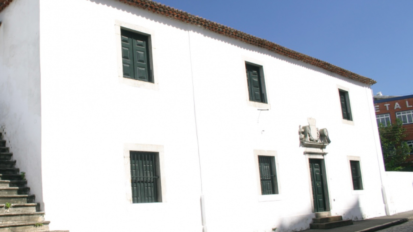 Construção branca de dois andares com escada externa e janelas verdes, combinando arquitetura tradicional com elementos decorativos em ambiente urbano