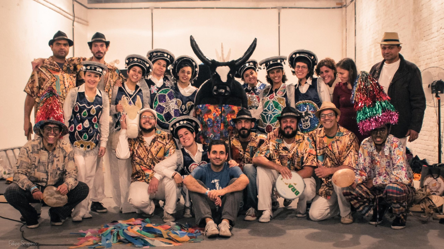 Grupo de artistas posa em conjunto, vestidos com trajes coloridos e tradicionais de bumba meu boi, ao centro uma figura de boi decorada