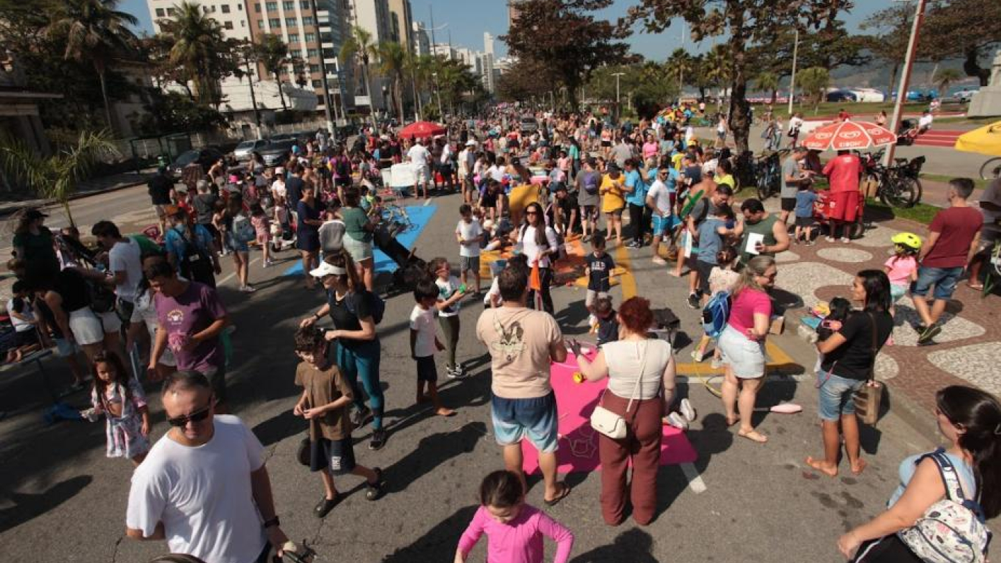 A imagem mostra uma grande concentração de pessoas participando de um evento ao ar livre em uma avenida à beira-mar, em um dia ensolarado. O local está fechado para carros e repleto de famílias, adultos e crianças, que caminham, andam de bicicleta e participam de atividades recreativas. Há mesas, brinquedos e áreas delimitadas por tapetes coloridos no chão, indicando ações interativas e lúdicas. Ao fundo, é possível ver prédios residenciais, coqueiros e parte da ciclovia à beira da praia.