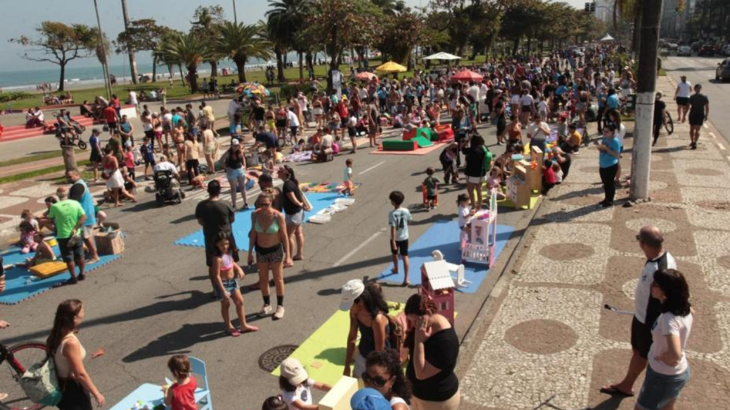 A imagem mostra um evento ao ar livre realizado na orla de Santos, em um dia ensolarado. A avenida à beira-mar foi fechada para veículos e está tomada por pessoas de todas as idades, especialmente famílias com crianças. Há diversas atividades recreativas espalhadas pela via, como jogos, brinquedos, oficinas e espaços coloridos montados com tatames e estruturas de papelão.
