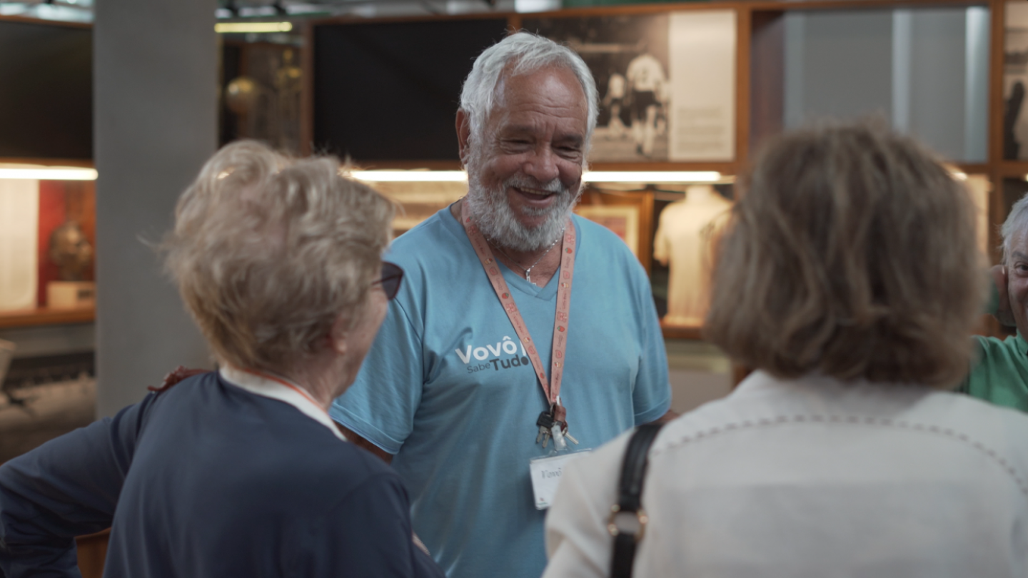 A cena mostra um grupo de pessoas em um ambiente interno, possivelmente um museu ou centro cultural, já que há vitrines e painéis com objetos e fotografias ao fundo. No centro da imagem está um senhor de barba e cabelos brancos, sorridente, usando uma camiseta azul clara com a inscrição “Vovô Sabe Tudo”. Ele tem um crachá pendurado no pescoço, preso a uma fita cor-de-rosa, e carrega um chaveiro.