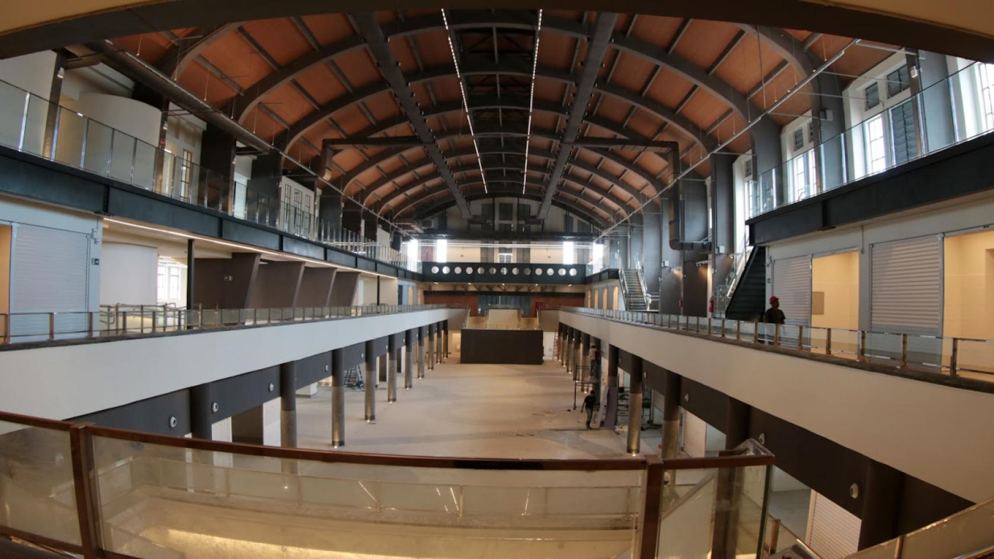 Esta é uma imagem panorâmica de um grande e espaçoso interior de mercado ou salão em reforma ou construção, com um teto abobadado de estrutura metálica aparente e áreas de mezanino em ambos os lados, com colunas e pisos limpos