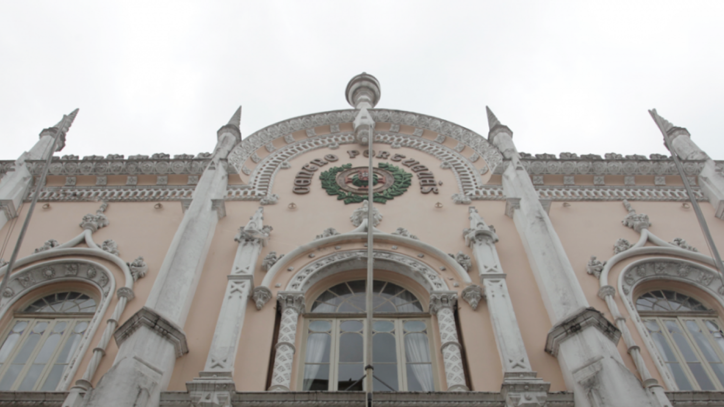 Vista em contraponto da fachada neogótica em tons de bege e branco do Centro Português, com três grandes janelas em arco e pináculos pontiagudos contra um céu claro. No centro superior, o emblema da instituição está gravado sob a balaustrada decorada.