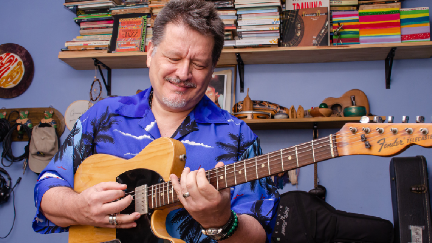 O músico Alexandre Birkett sorri enquanto toca uma guitarra Fender Telecaster em seu estúdio, que tem prateleiras cheias de livros e objetos de arte no fundo azul