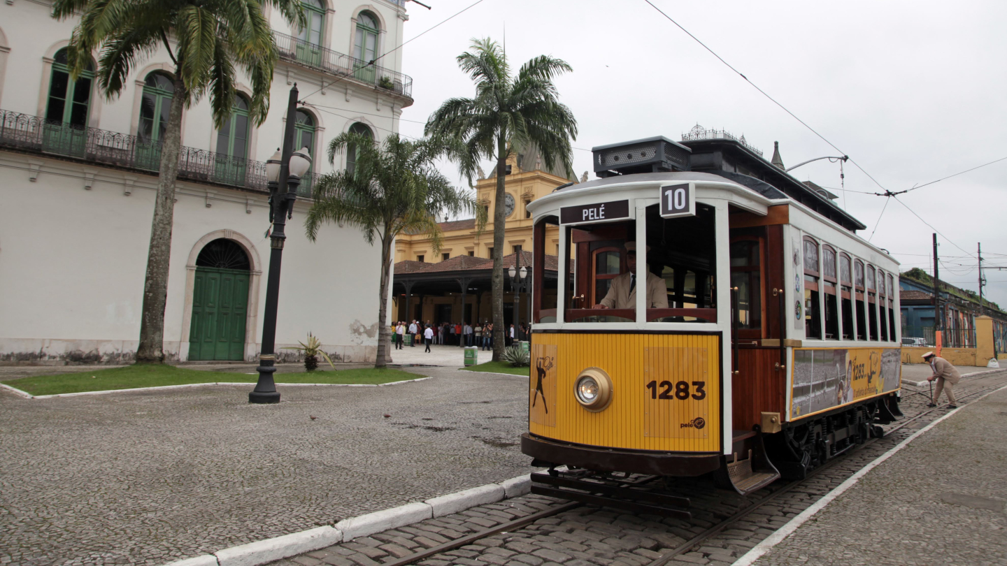 Imagem mostra um bonde histórico amarelo e branco, identificado como “Pelé”, circulando por uma rua de paralelepípedos no Centro Histórico de Santos. Ao fundo, aparecem prédios antigos, palmeiras e pedestres próximos à estação.