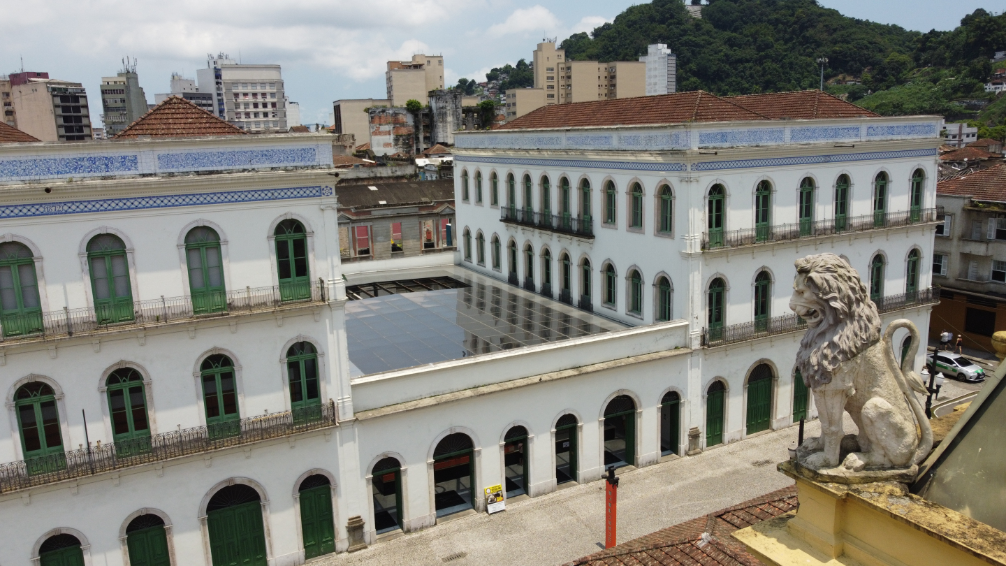 Uma vista panorâmica da arquitetura histórica de Santos, mostrando dois edifícios brancos de estilo colonial com janelas em arco e caixilhos verdes, um pátio central com cobertura de vidro e uma escultura de leão em primeiro plano à direita